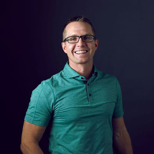 Man with glasses smiling in a green polo shirt against a dark background.