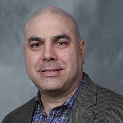 A professional headshot of a bald man with dark eyes wearing a grey jacket.