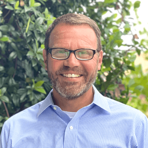 Smiling man with glasses and beard wearing a blue shirt against a green foliage background.