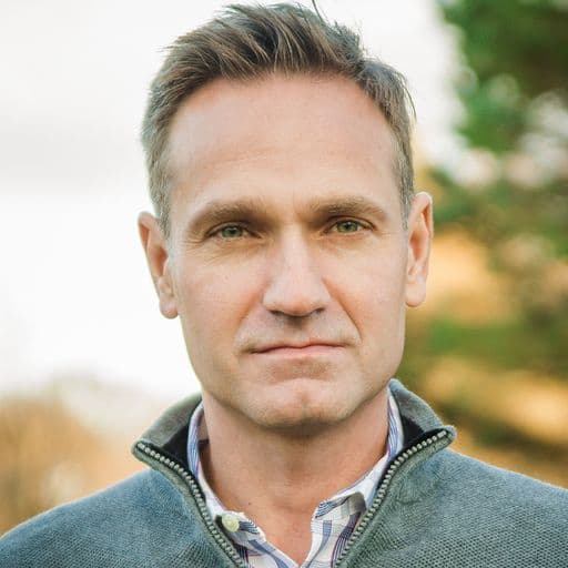 Professional headshot of a man with short brown hair wearing a green jacket outdoors.