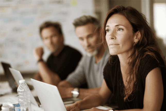 Attentive woman at a business meeting with laptops and colleagues in background.