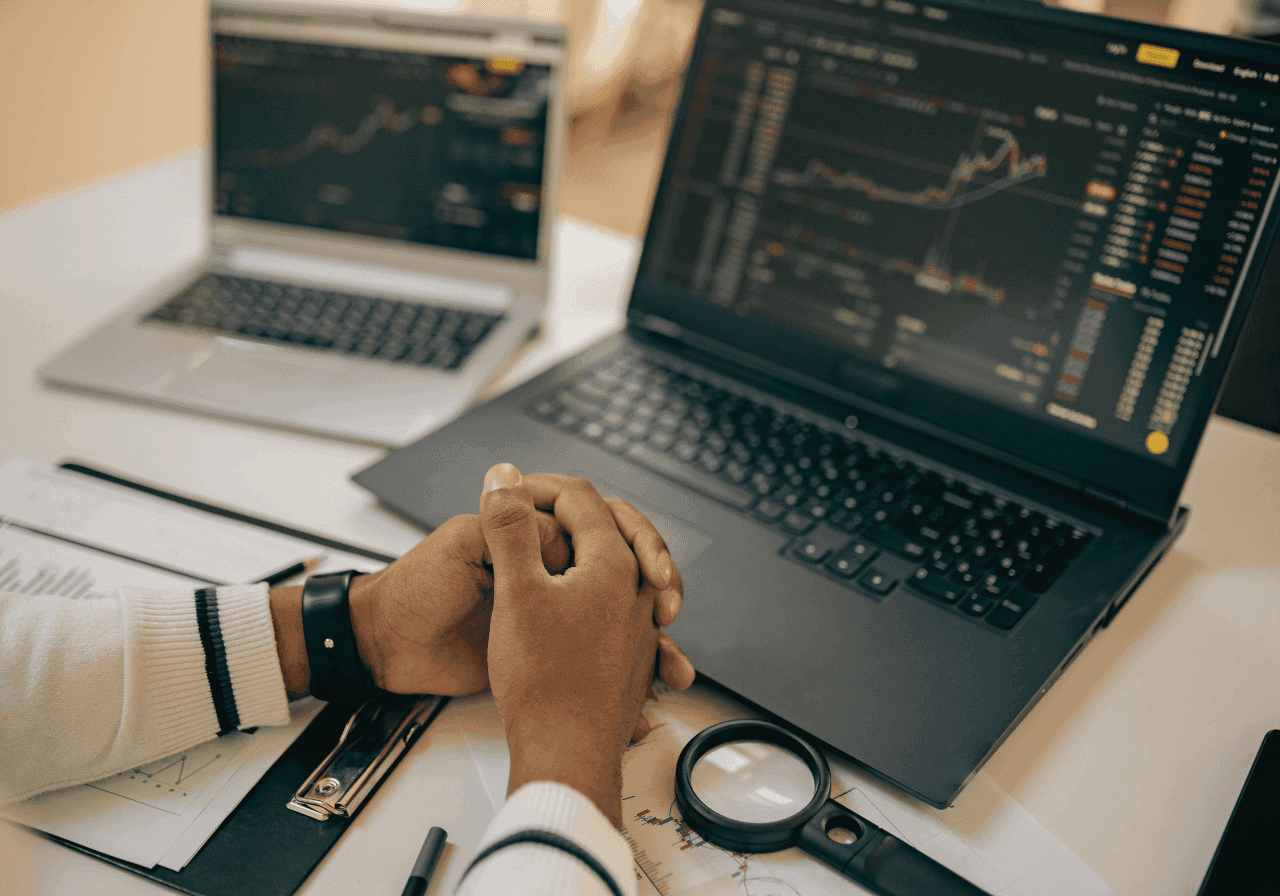 Hands clasped over a desk with two laptops displaying financial market charts and data.