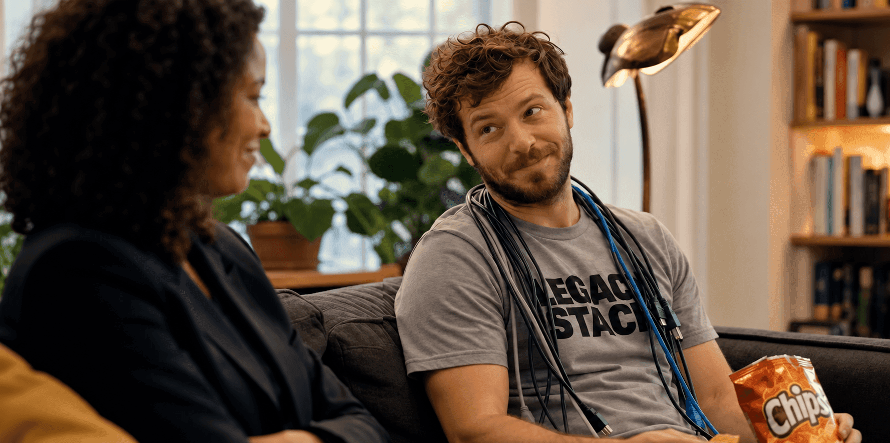 A man on a couch smiling at a woman, holding a bag of chips.