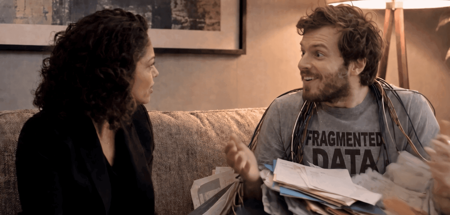 A woman listens as a man in a 'FRAGMENTED DATA' shirt gestures with papers.