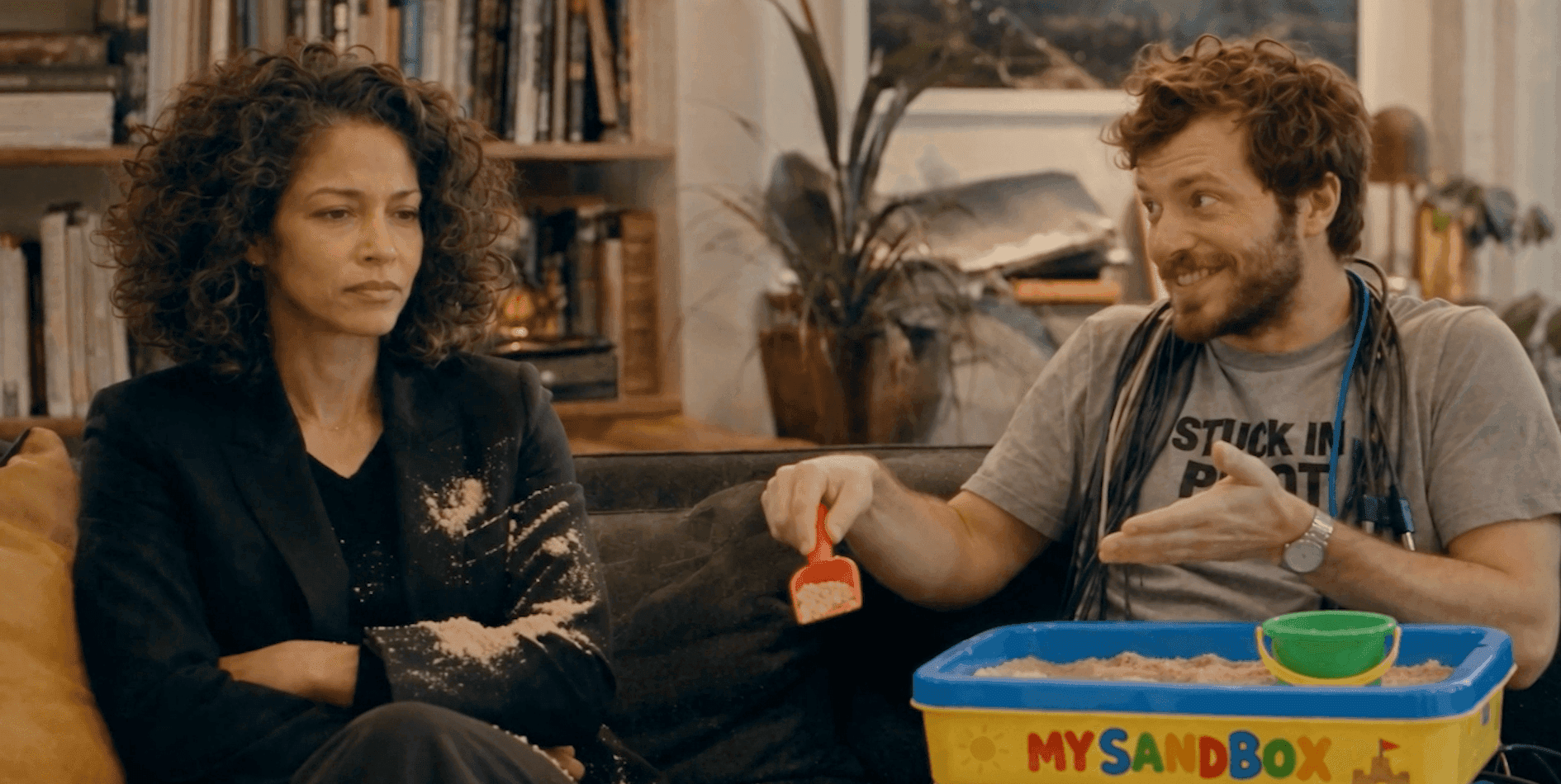 A man with a toy sandbox talks to an unamused woman covered in sand.