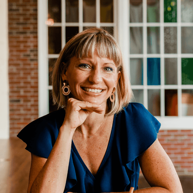 Smiling woman with blonde hair in a blue top, hand resting on her chin.