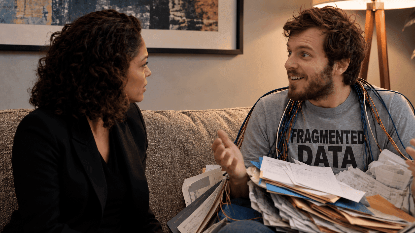 Man in 'FRAGMENTED DATA' shirt talks to woman while holding disorganized papers.