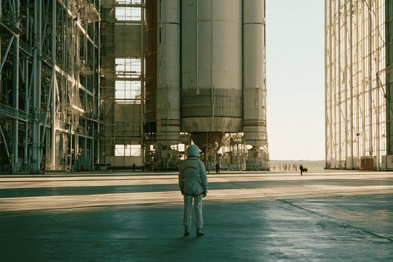 Person in white suit stands before a giant rocket inside a vast industrial hangar.